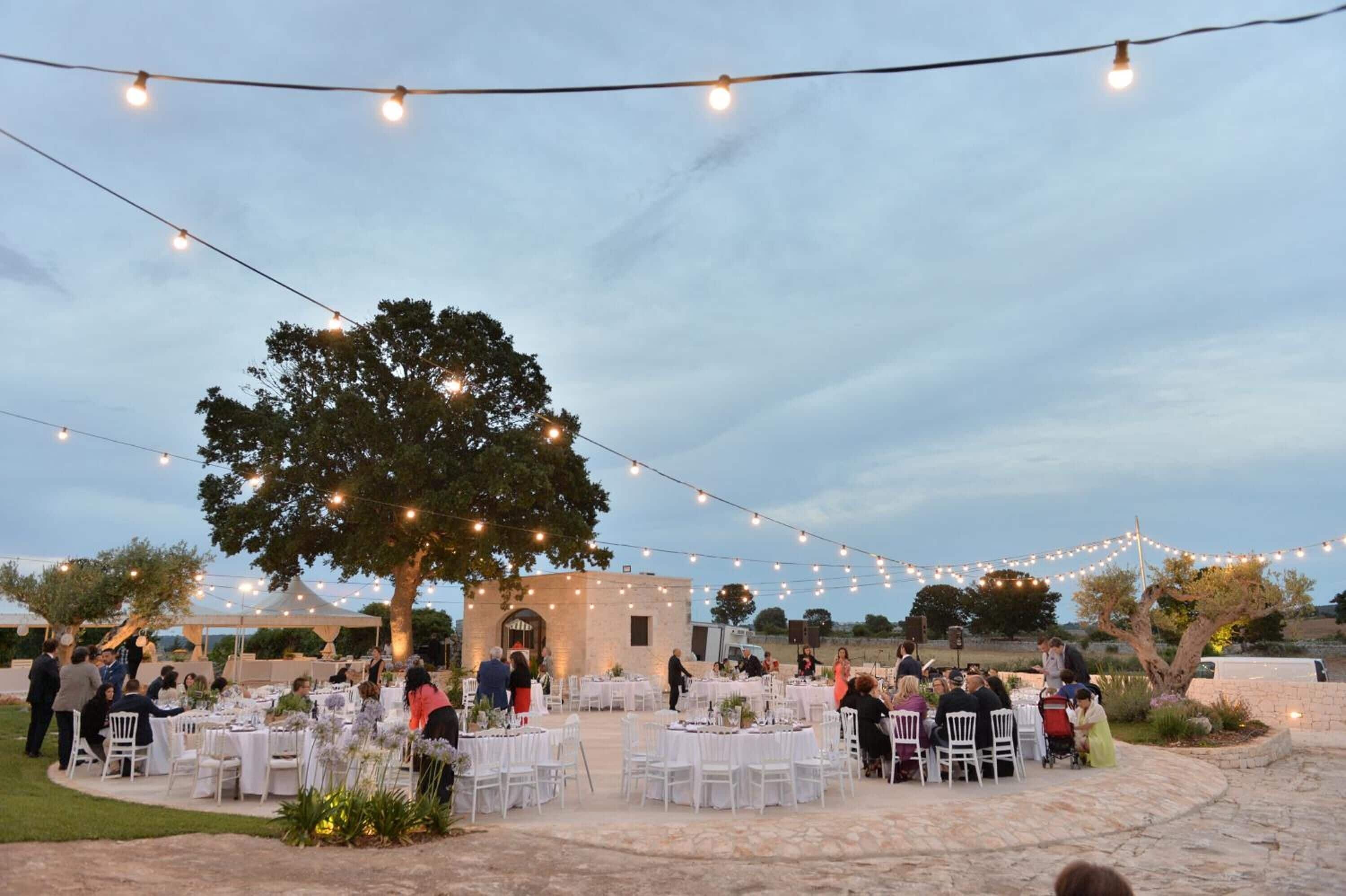 An outdoor event with round tables and chairs, string lights, and a large tree.
