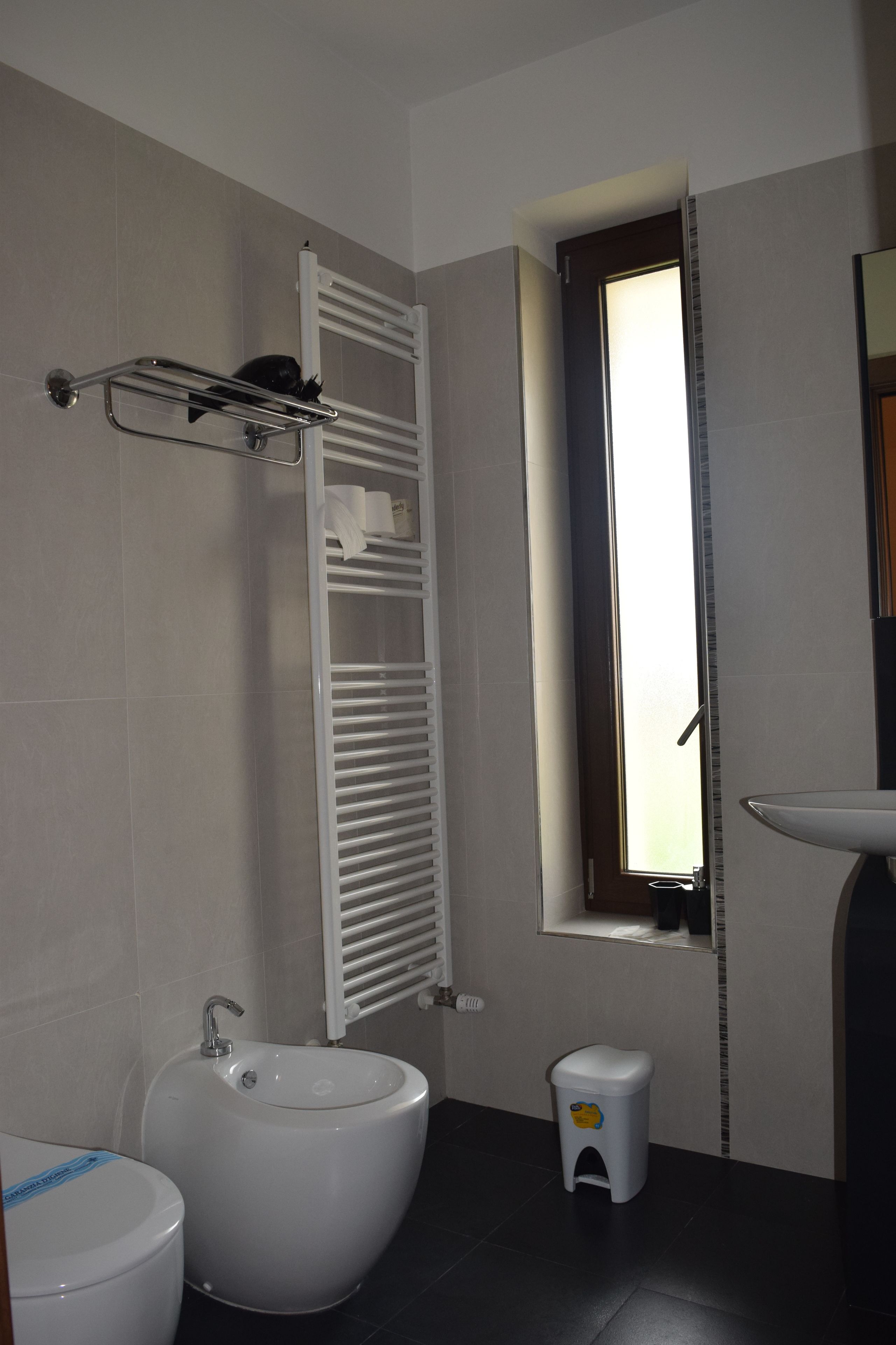 A bathroom with a bidet, a small stool, a window, and a towel rack.