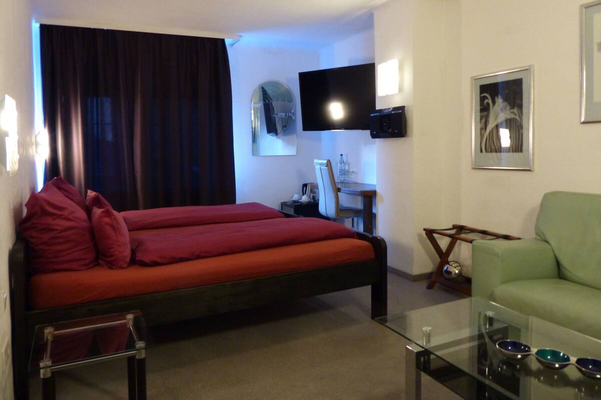 A hotel room with a bed, a green armchair, a glass coffee table, a desk with a chair, a flat-screen TV mounted on the wall, and a framed picture.