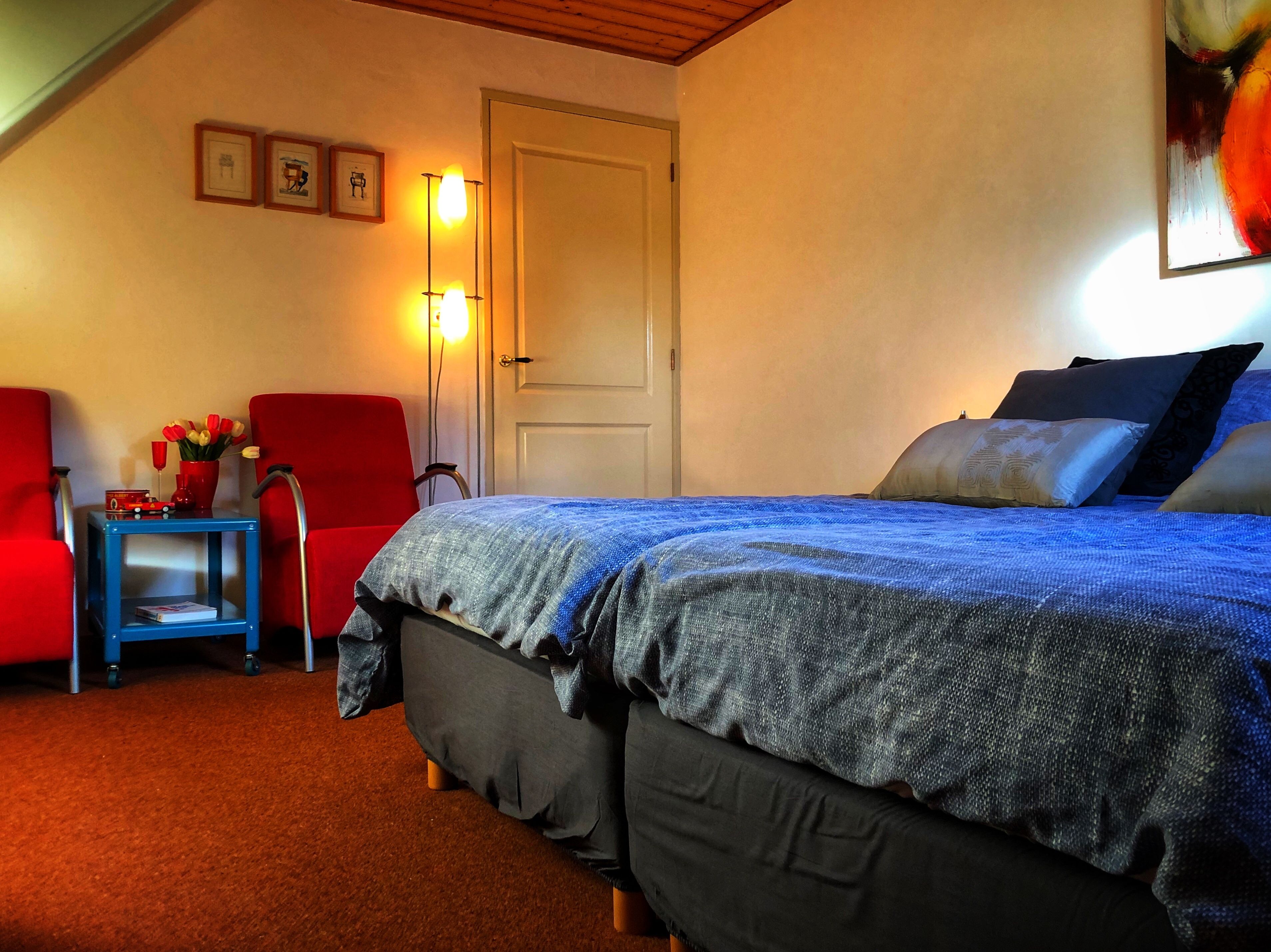 A bedroom with a bed, a red chair, a small table with a vase of flowers, and a door.