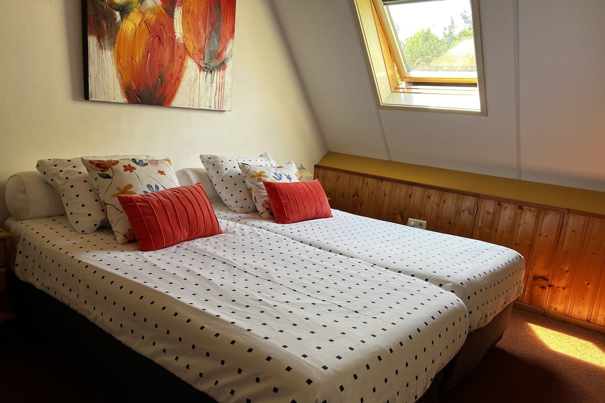 Une chambre avec deux lits, chacun recouvert d’une courtepointe blanche ornée de pois noirs, et agrémentée d’oreillers rouges. Un tableau est accroché au mur, au-dessus des lits, et une fenêtre de toit se trouve sur le côté droit.