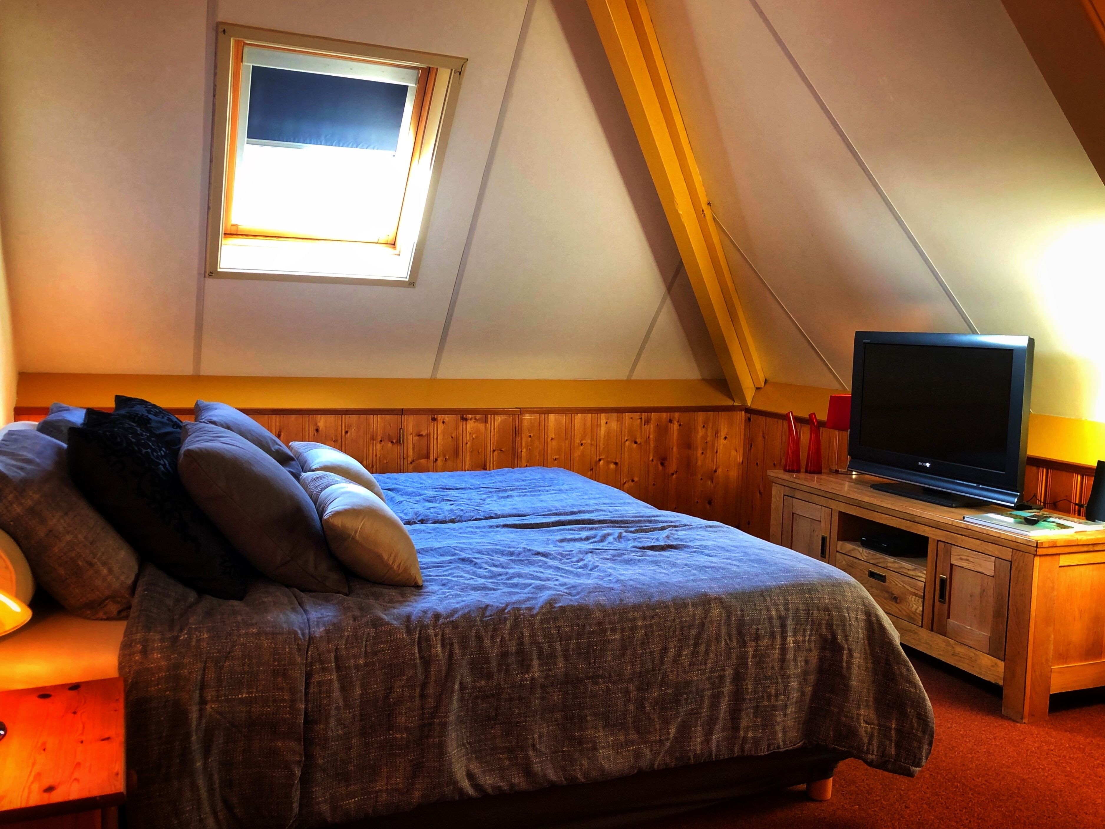 A bedroom with a sloped ceiling, a bed with pillows, a TV on a wooden stand, and a window with blinds.
