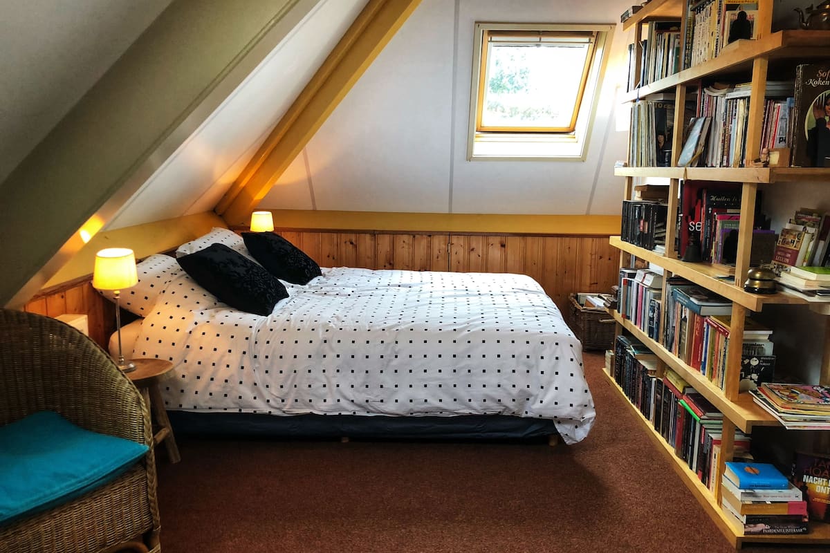 Une chambre mansardée chaleureuse, avec un plafond en pente, une petite lucarne, un lit recouvert d’une couette à motifs noirs et blancs, deux oreillers noirs, une chaise en osier et une bibliothèque remplie de livres.