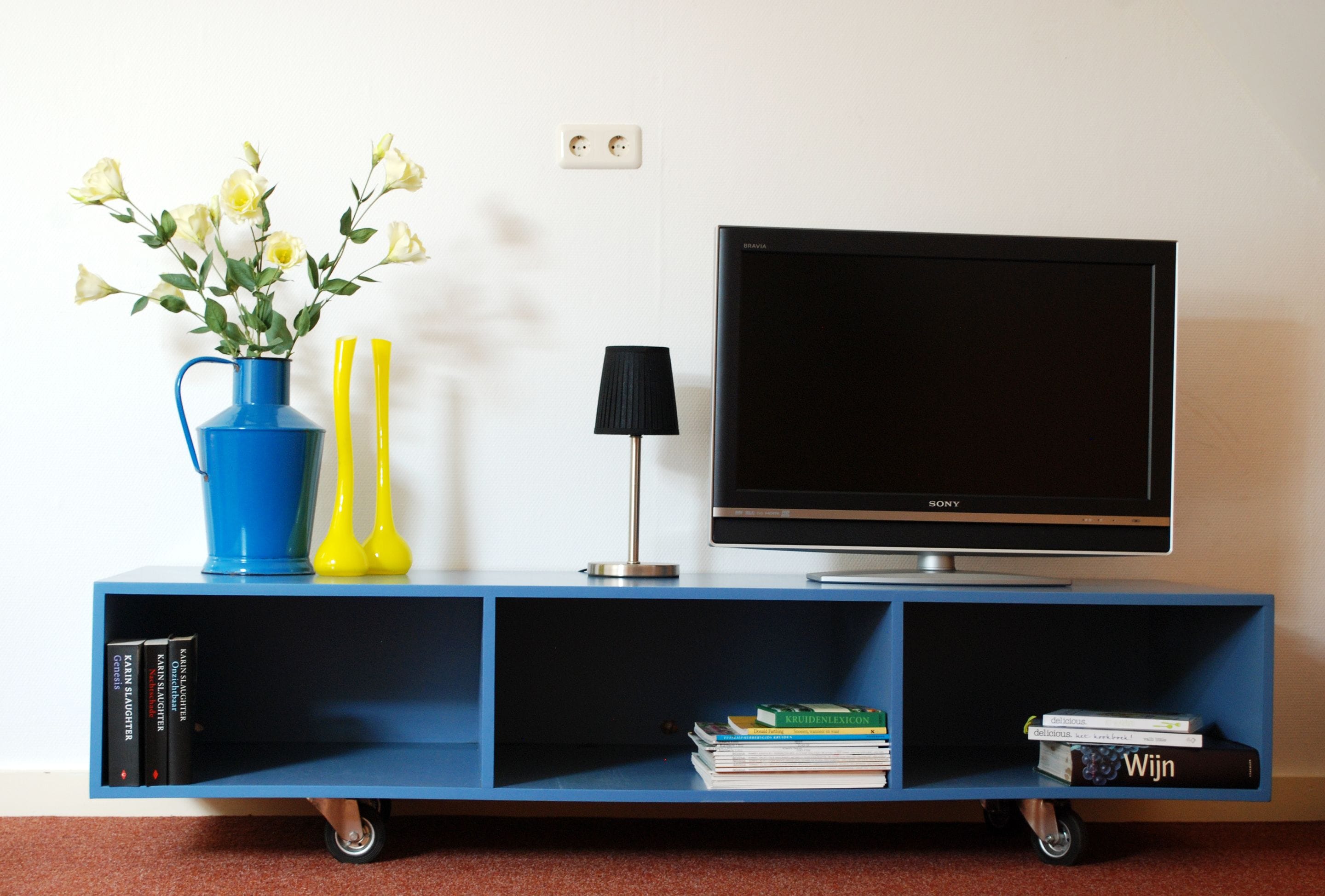 Een blauwe entertainmentkast met een flatscreen televisie, een vaas met bloemen, een lamp en boeken.