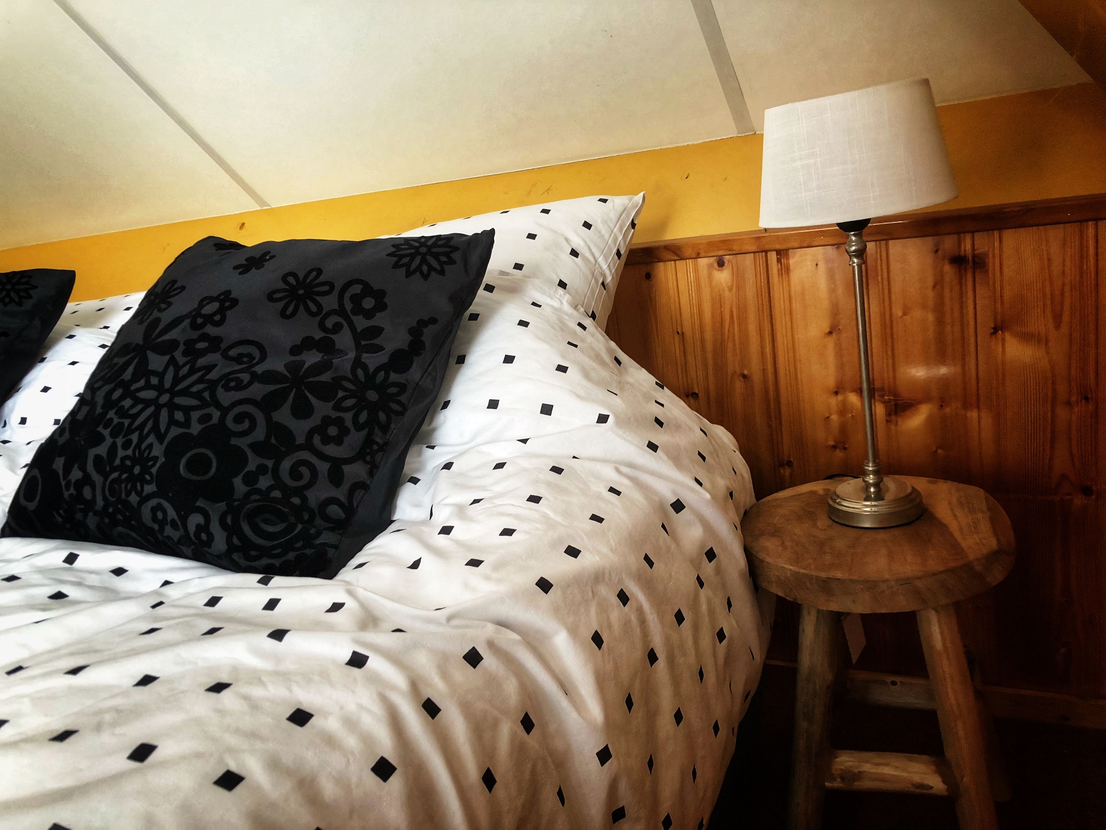 A bed with patterned pillows, a wooden nightstand with a lamp, and a yellow wall.