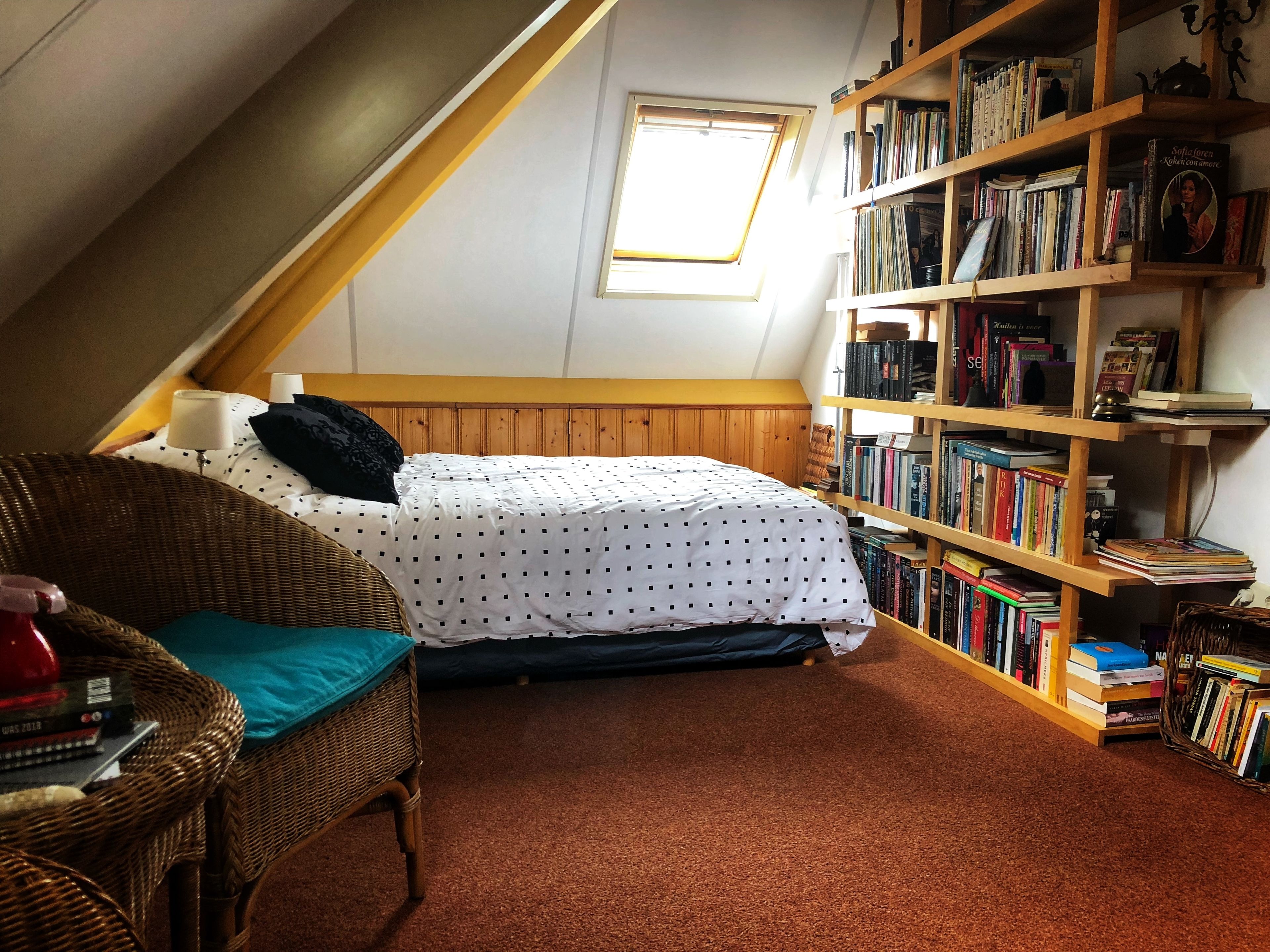 A cozy attic bedroom with a sloped ceiling, a wooden bookshelf filled with books, a wicker chair, and a neatly made bed.
