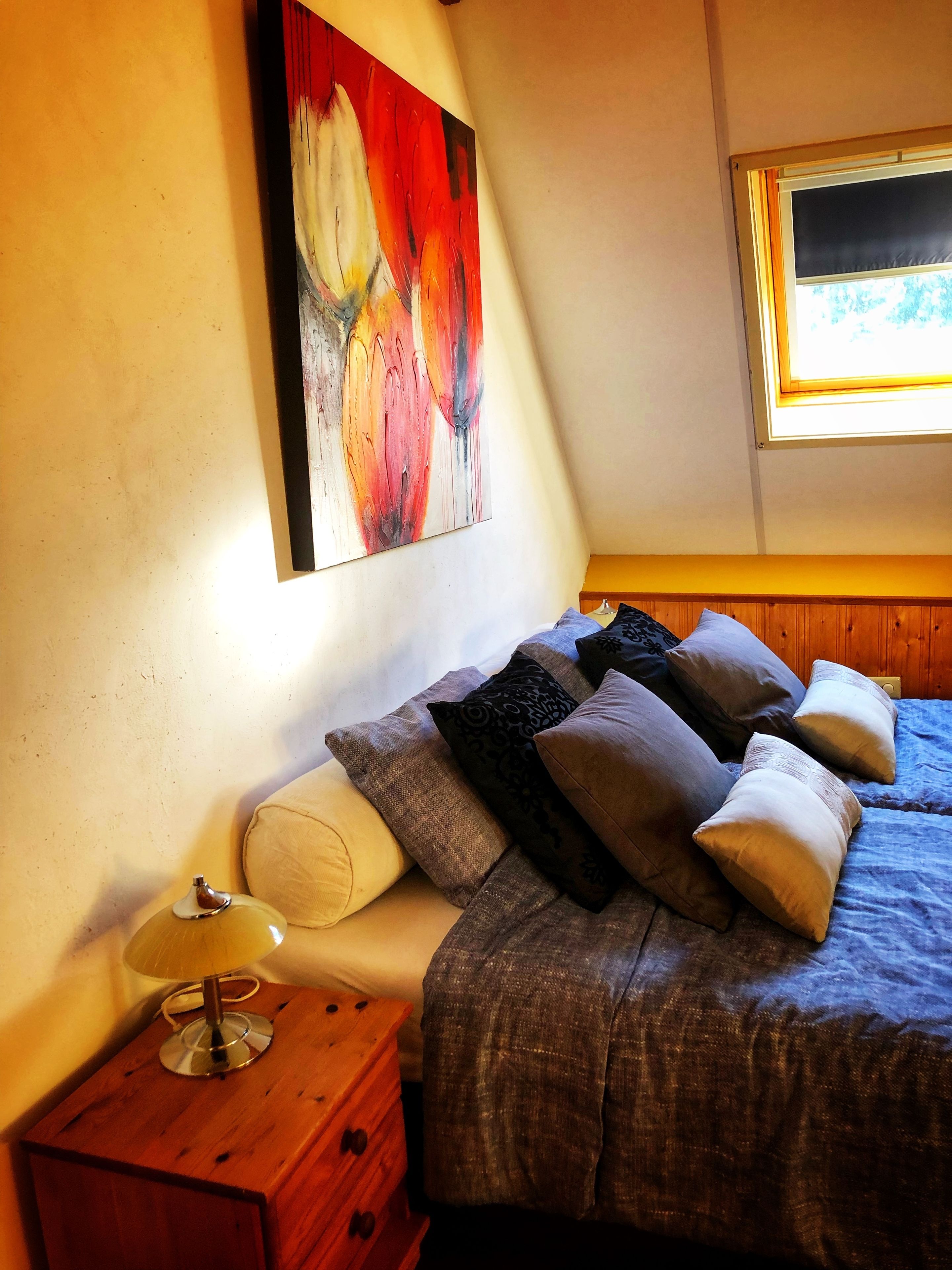 Un dormitorio con cama, almohadas, una mesita de noche con lámpara, un aparador de madera y un cuadro en la pared.