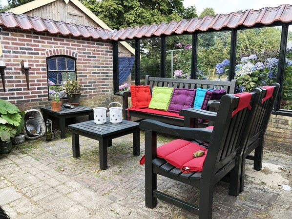 A patio area with a brick wall, a wooden bench with colorful cushions, a small table with a potted plant, and a metal chair. - B&B Zandhuizen (Zandhuizen)
