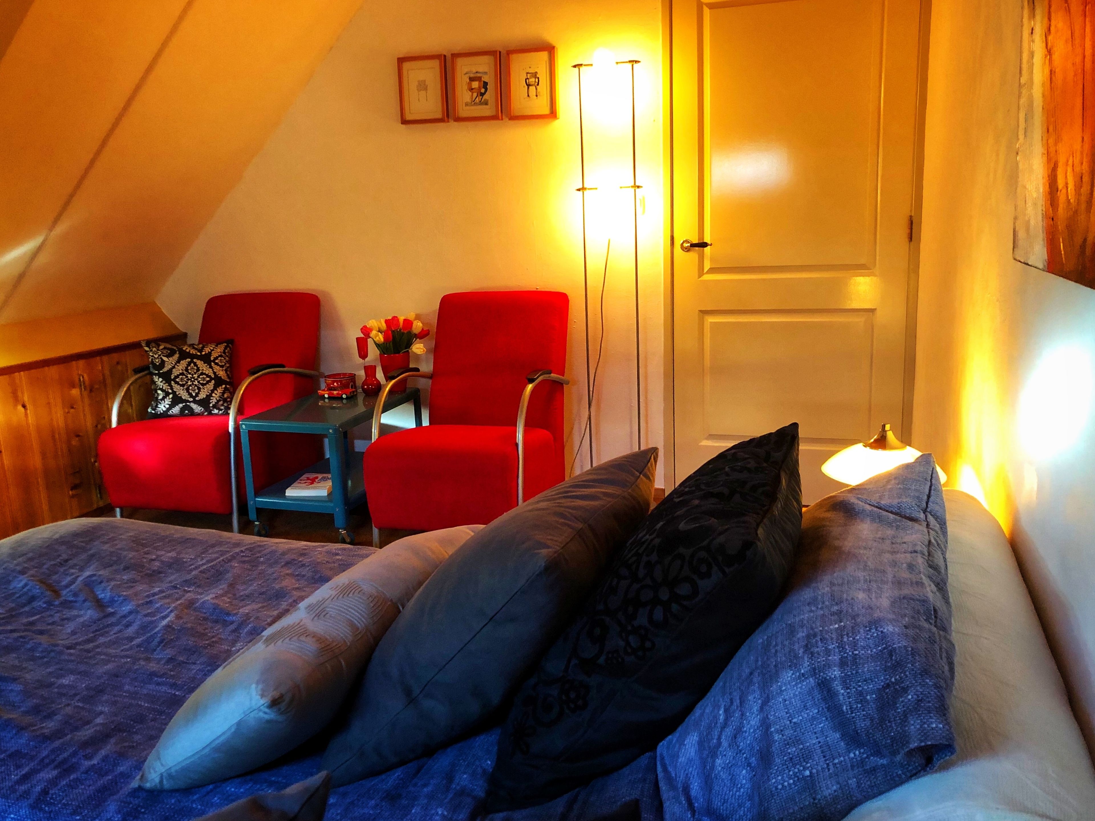 Um quarto acolhedor com teto inclinado, uma cama com almofadas estampadas, duas cadeiras vermelhas, uma mesa pequena com um vaso de flores e uma porta com interruptor de luz.