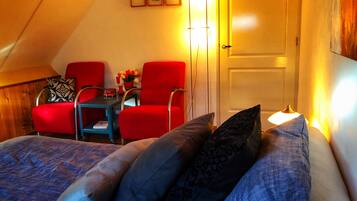 Una stanza accogliente con soffitto a falde, un letto con cuscini decorati, due poltrone rosse, un tavolino con un vaso di fiori e una porta con interruttore della luce.