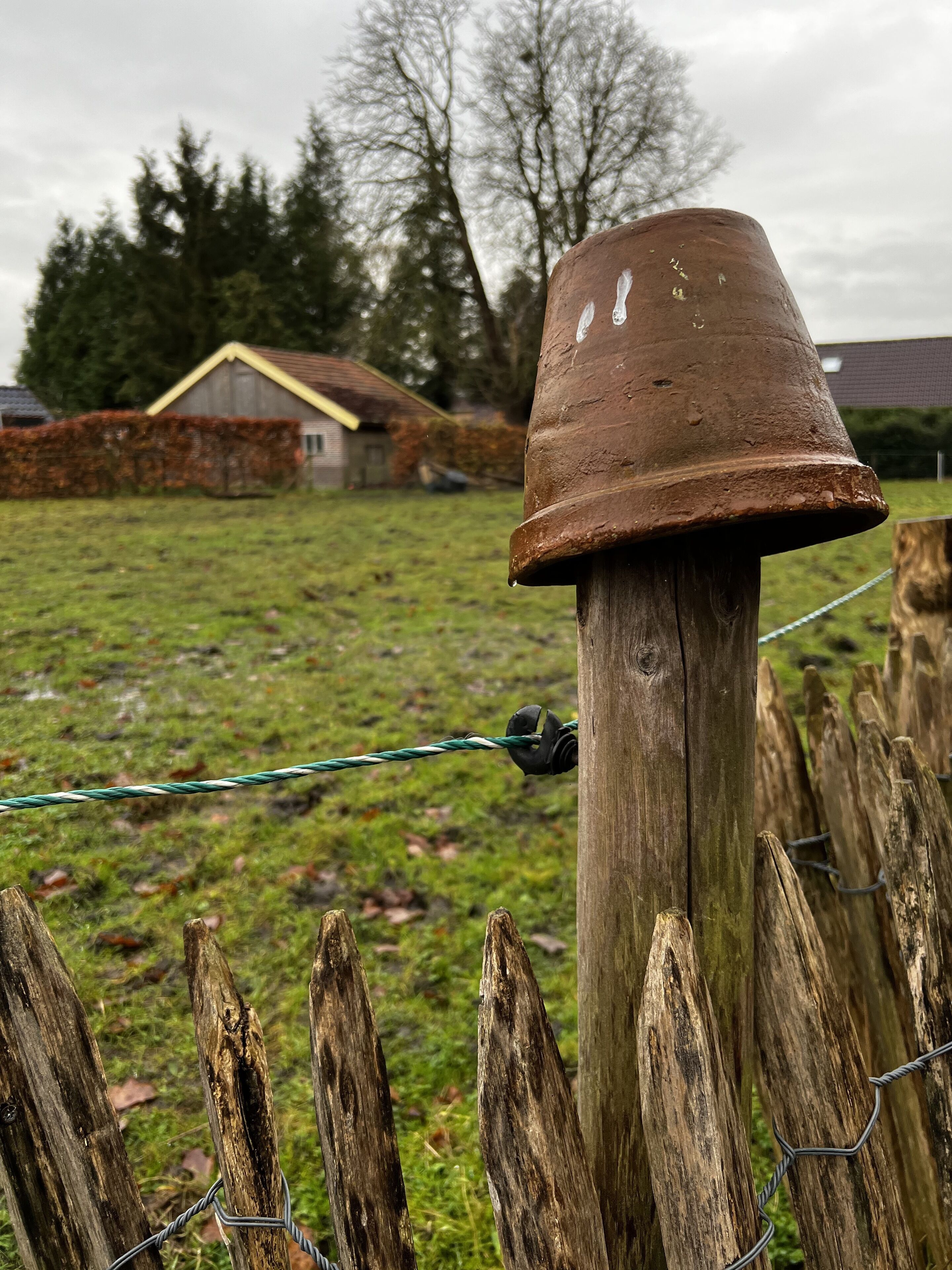 A rustic wooden fence with a metal gate, a weathered brown pot on top, and a grassy field with a house in the background.