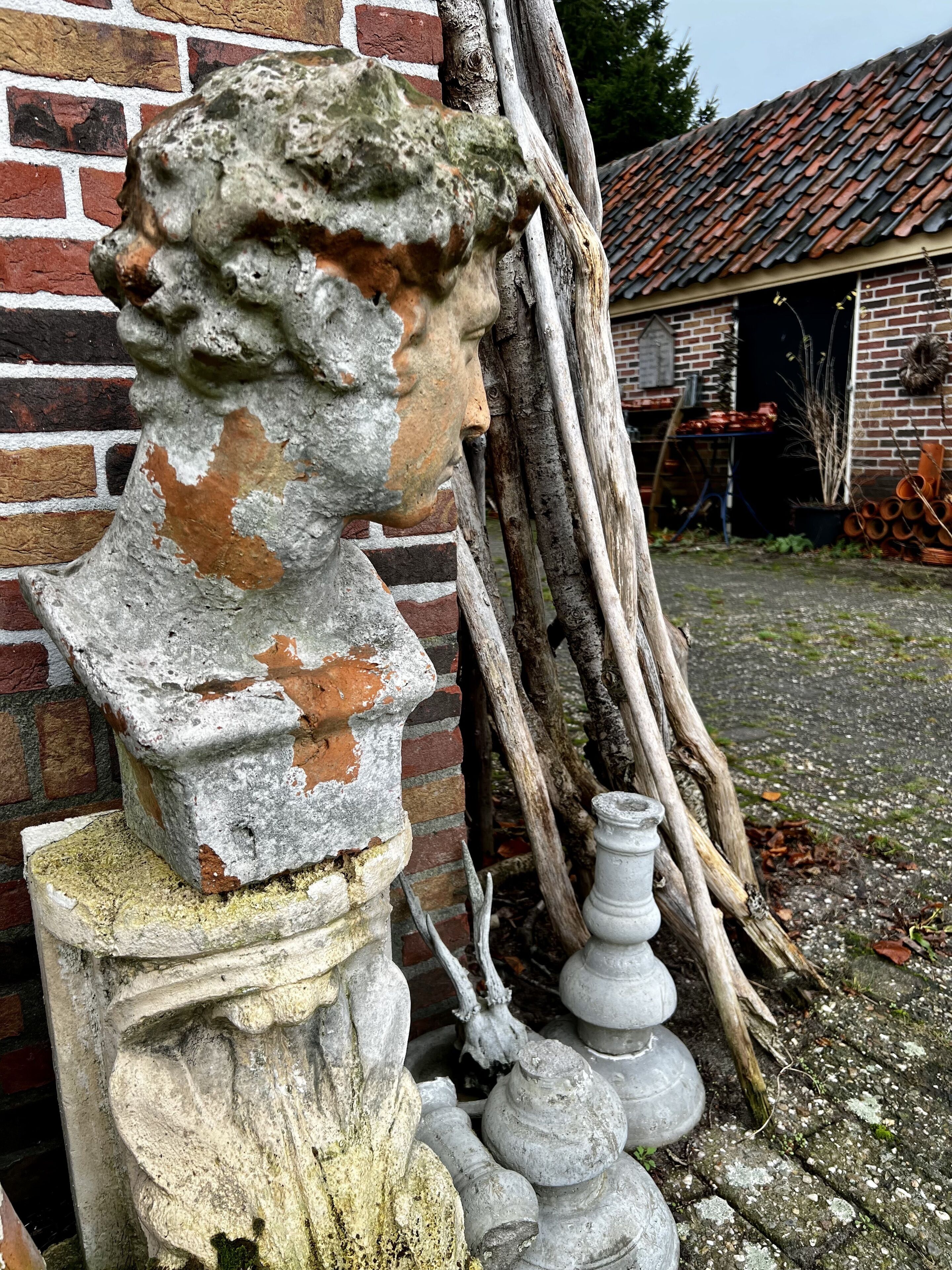 Een verweerd bustebeeld tegen een bakstenen muur, met een rustiek gebouw en tuingereedschap op de achtergrond.