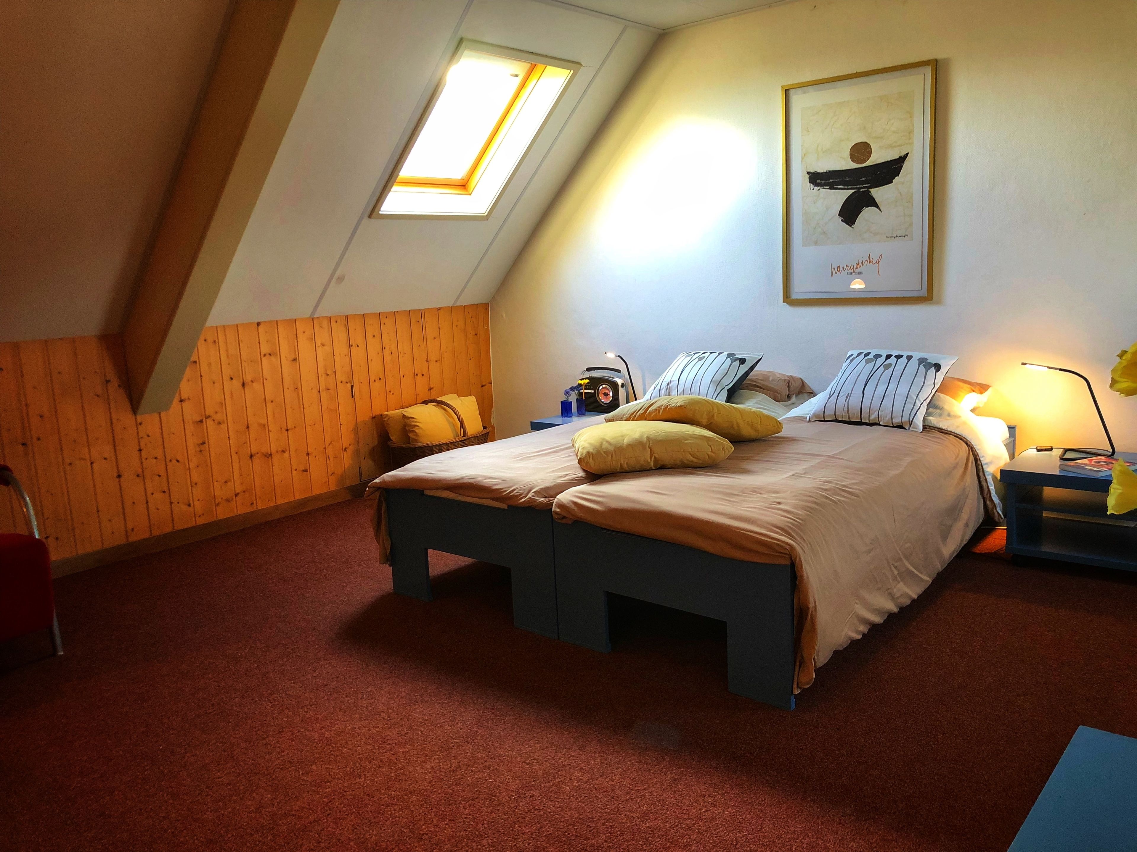 Un dormitorio con techo inclinado, una cama con almohadas amarillas, una obra de arte enmarcada en la pared y una mesita de noche con una lámpara.
