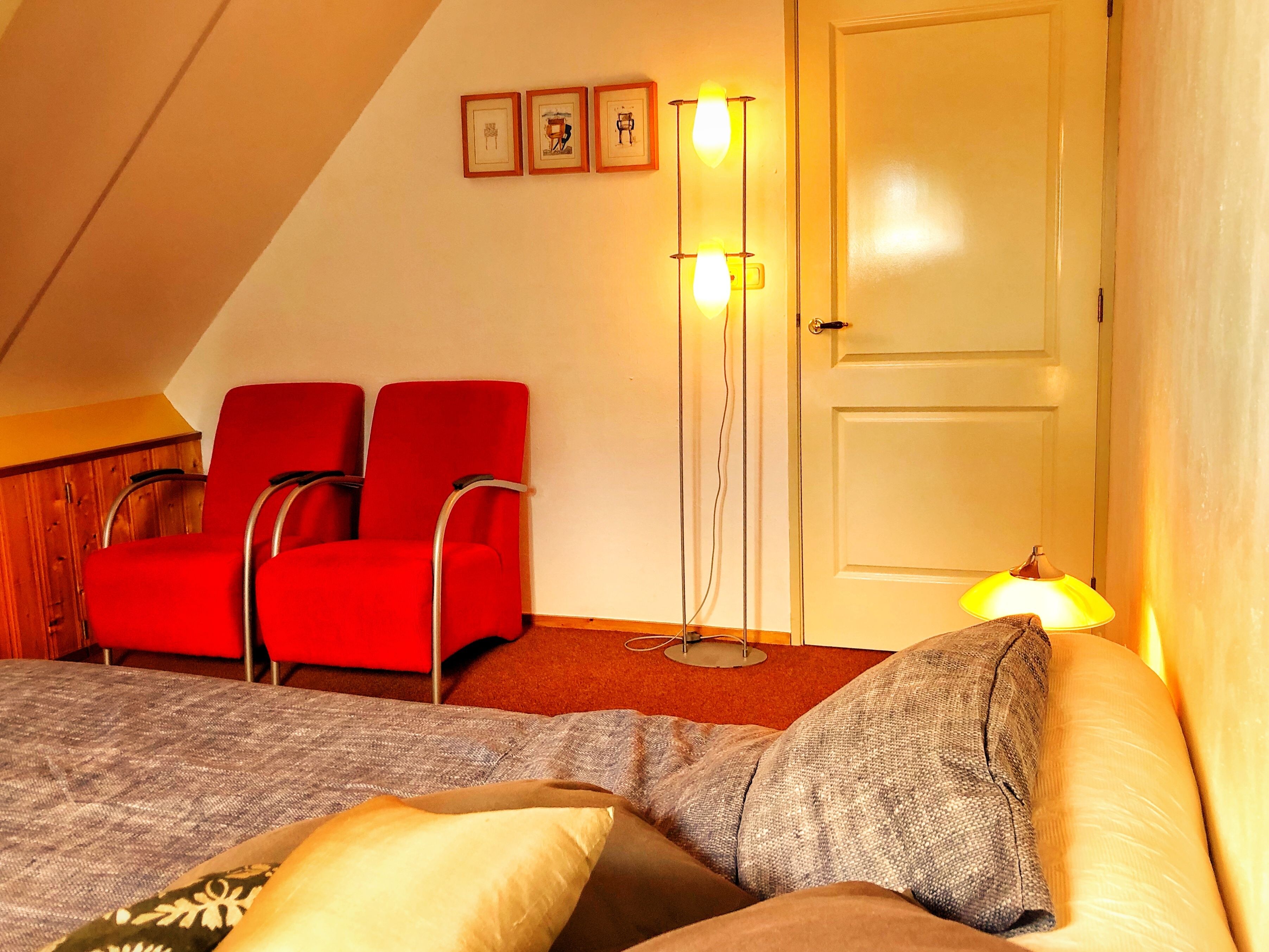 Een kamer met een bed, twee rode stoelen, een vloerlamp en een deur.