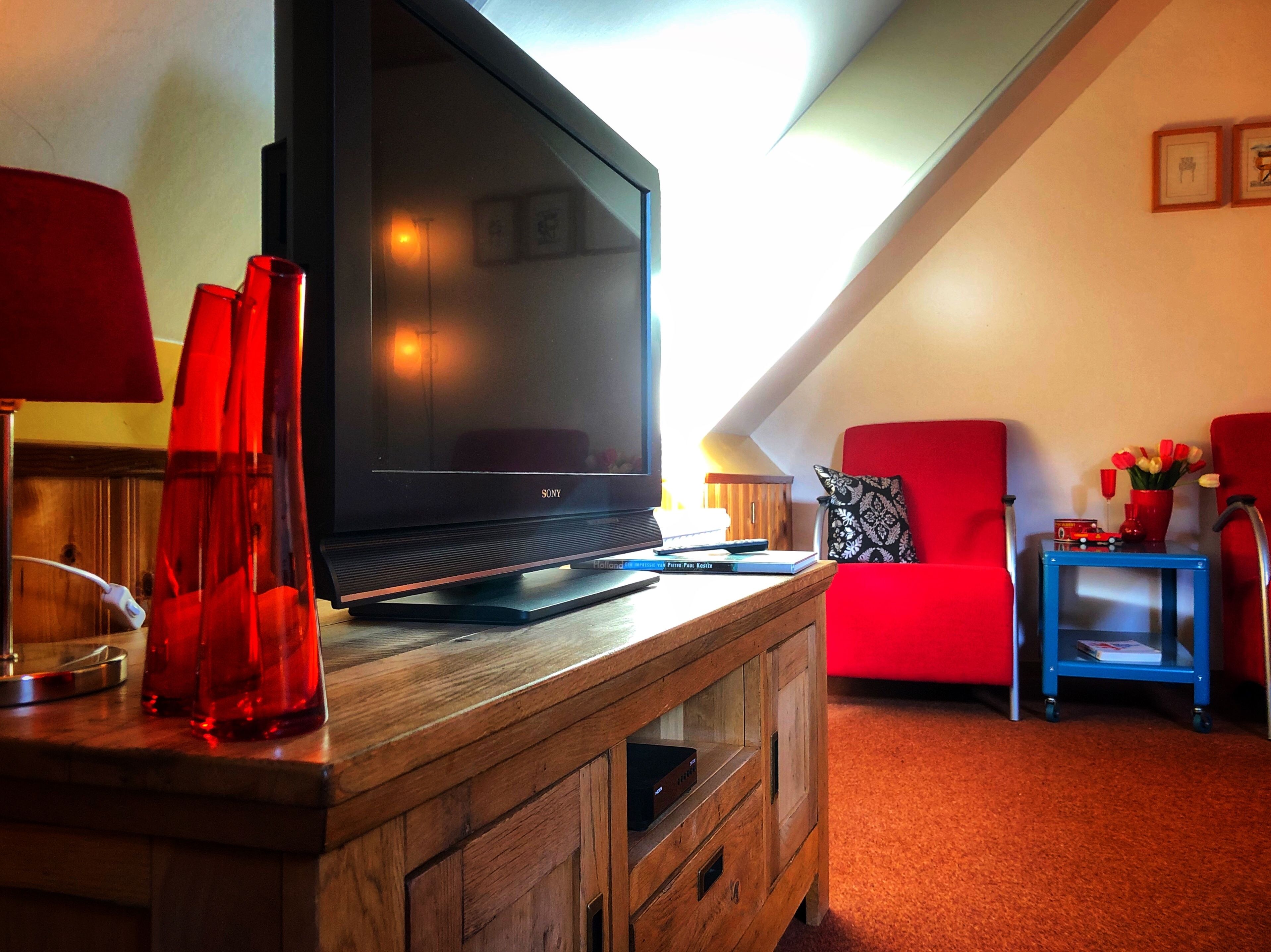 A modern living room with a flat-screen TV on a wooden stand, a red armchair, a small side table with a red vase and a toy train, and a red lamp on a wooden cabinet.