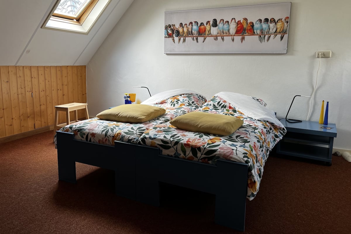 Une chambre avec un plafond en pente, un lit recouvert d’une literie à motifs floraux, deux oreillers, une petite table de chevet en bois, une lampe et un tableau représentant des oiseaux, accroché au mur.