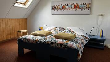 Un dormitorio con techo inclinado, una cama con ropa de cama floral, dos almohadas, una mesita de noche de madera y una lámpara, además de un cuadro de pájaros colgado en la pared.