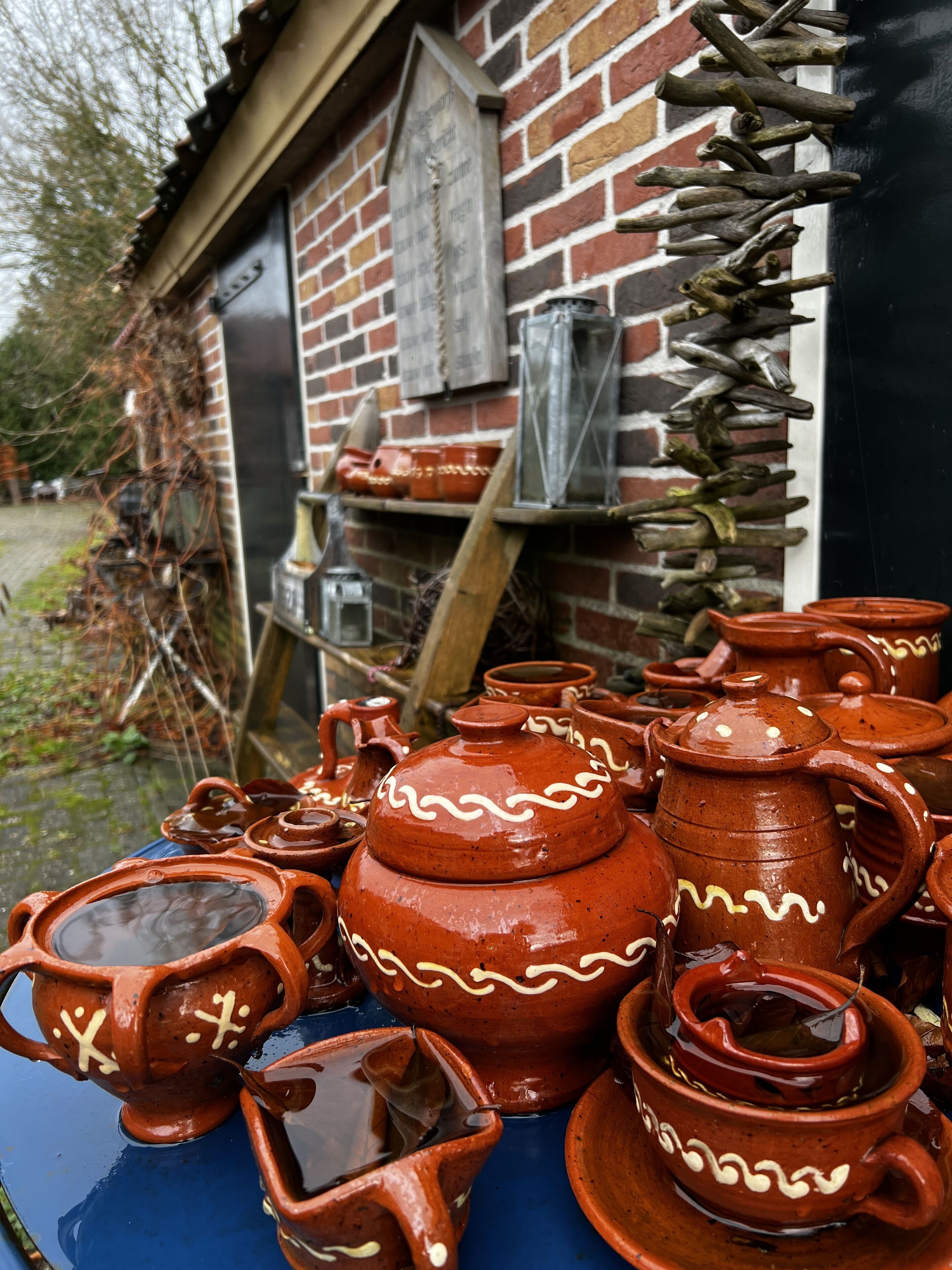 A collection of traditional clay pots and utensils displayed outside a brick building with a wooden ladder and a hanging vine.