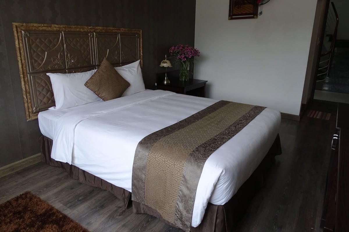 Una cama bien hecha con una colcha y almohadas estampadas, un cabecero de madera, una mesita de noche con una lámpara y flores, y un cuadro en la pared.