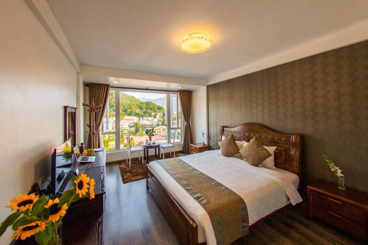 Habitación de hotel con una cama grande, un escritorio con una computadora portátil, una silla, vistas de un paisaje urbano a través de ventanas amplias y una lámpara en el techo.