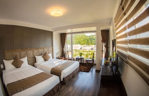 A hotel room with two beds, a small table, a TV, and a view of a city and mountains. - Gem Sapa Hotel (Sa Pa)