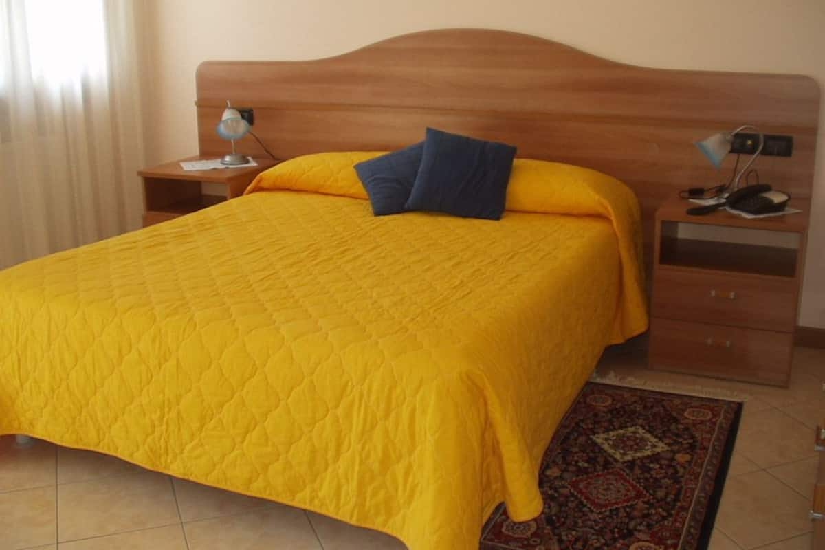 A hotel room with a wooden headboard, a yellow quilted bedspread, two pillows, a nightstand with a lamp, a drawer, and a patterned rug.