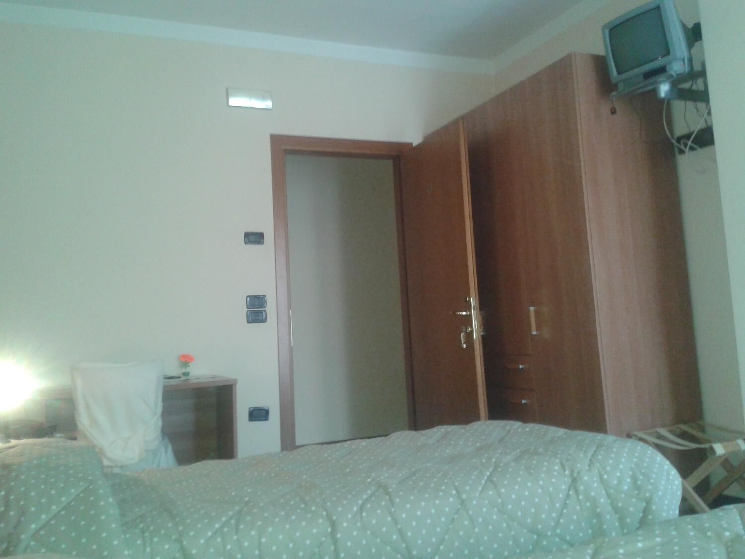 A bedroom with a bed, a wooden wardrobe, a small bedside table with a lamp, and a television mounted on the wall.