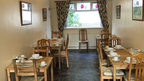 A dining area with wooden tables and chairs, a window with curtains, and a tiled floor. - The New Inn Guest House (Bridgend)