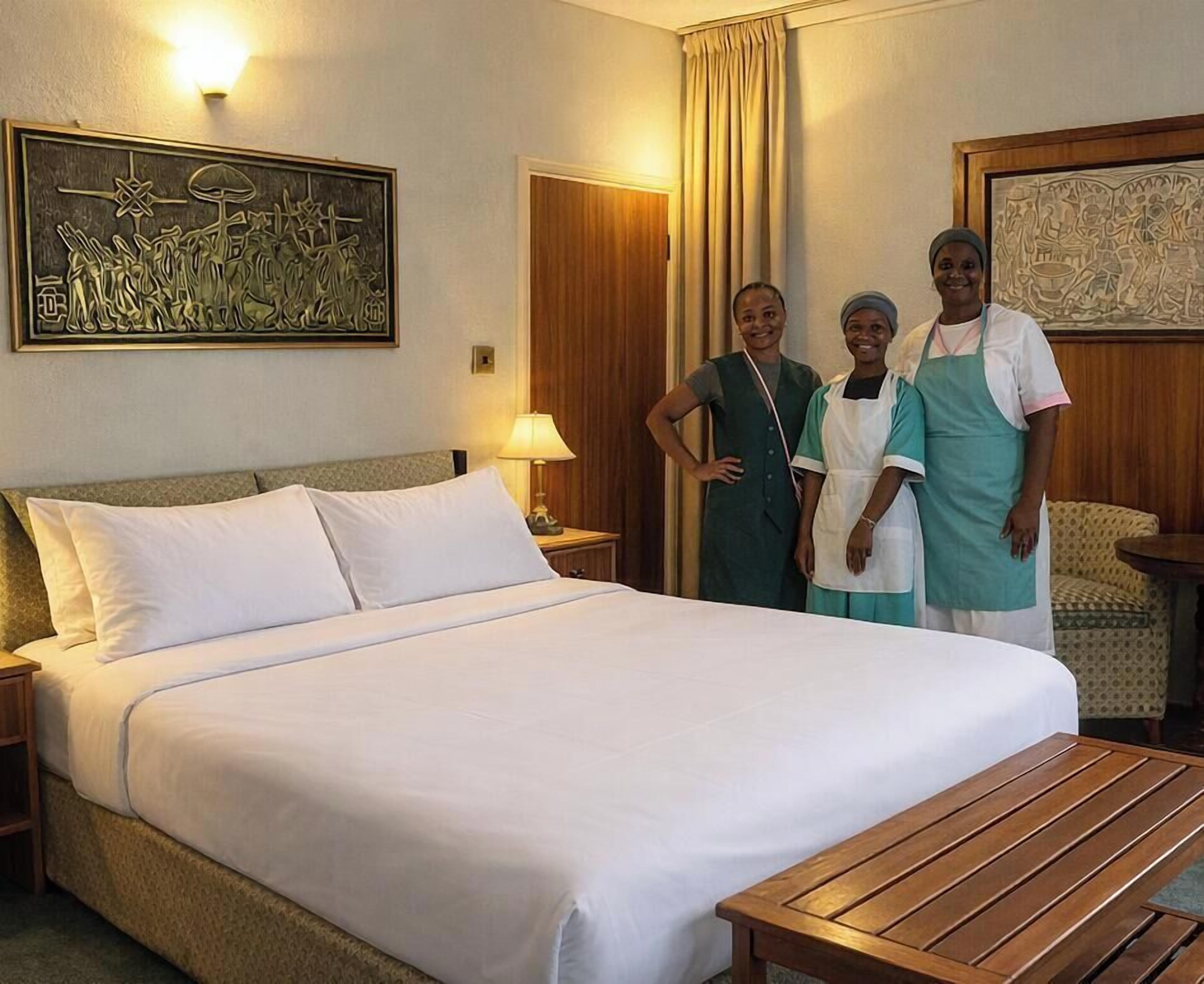 A hotel room with a large bed, a wooden nightstand, a lamp, a painting on the wall, and a wooden table.