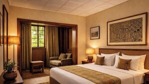 Habitación de hotel con una cama, dos mesitas de noche, una silla, una ventana con cortinas, una obra de arte en la pared y una planta en maceta.