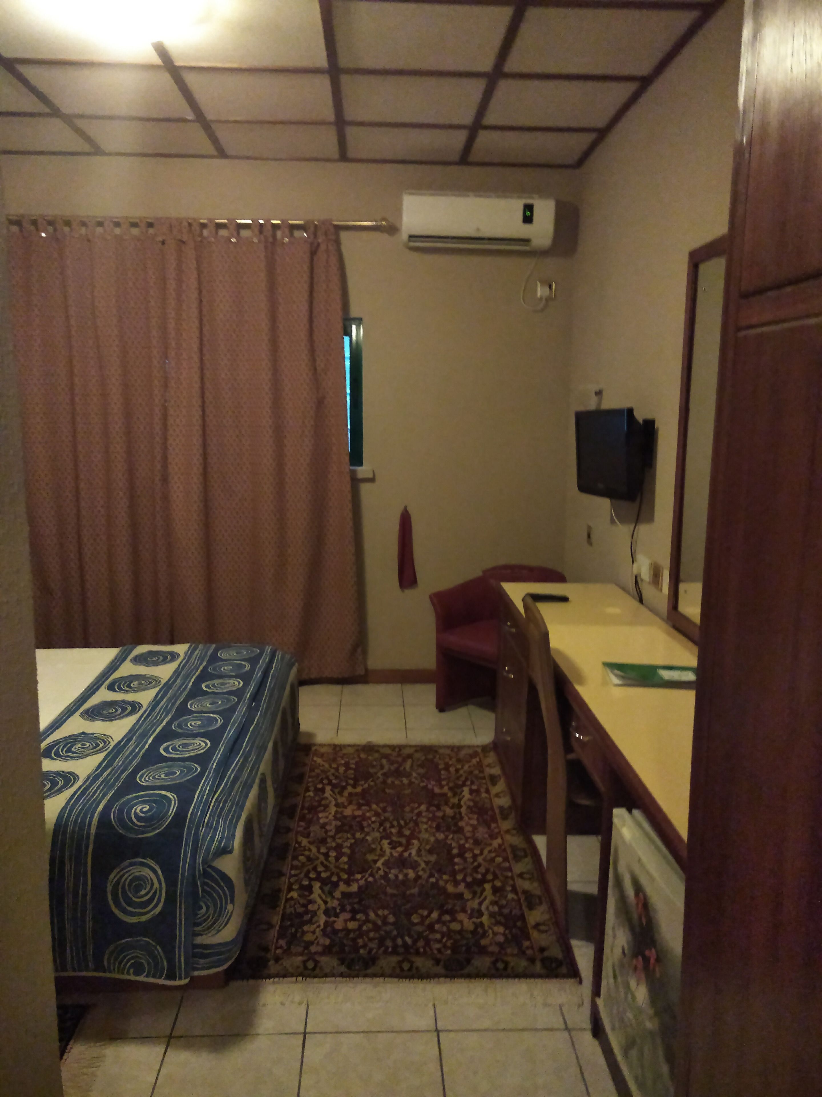 A small, dimly lit room with a bed, a desk, a chair, an air conditioner, and a television.