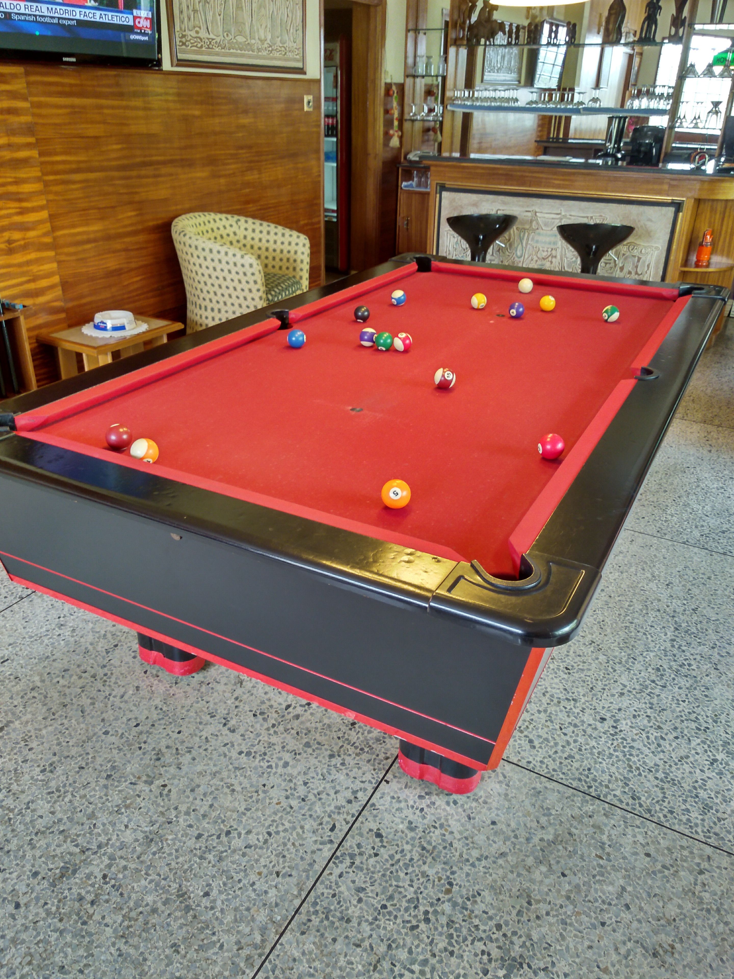 Una mesa de billar con bolas, un televisor mostrando un canal deportivo y una silla al fondo.