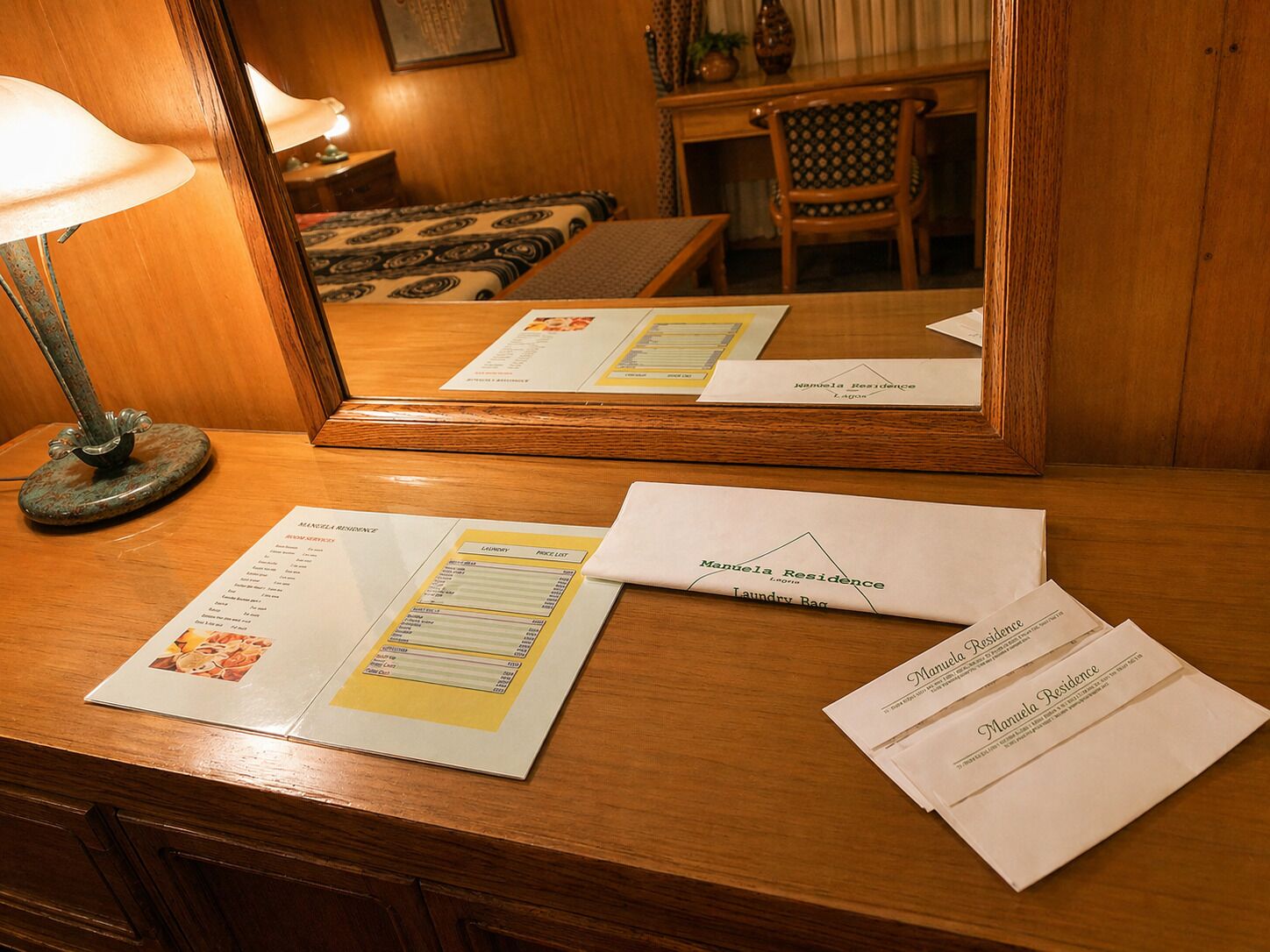A wooden desk with a mirror reflecting a bed, a lamp, and a chair. On the desk, there are printed materials including a menu and a laundry bag.
