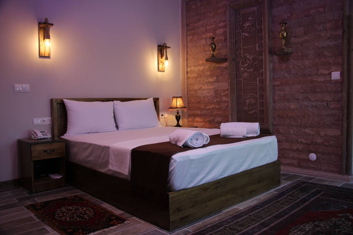 A neatly made bed with a wooden headboard, a nightstand with a phone, and a wall-mounted lamp.
