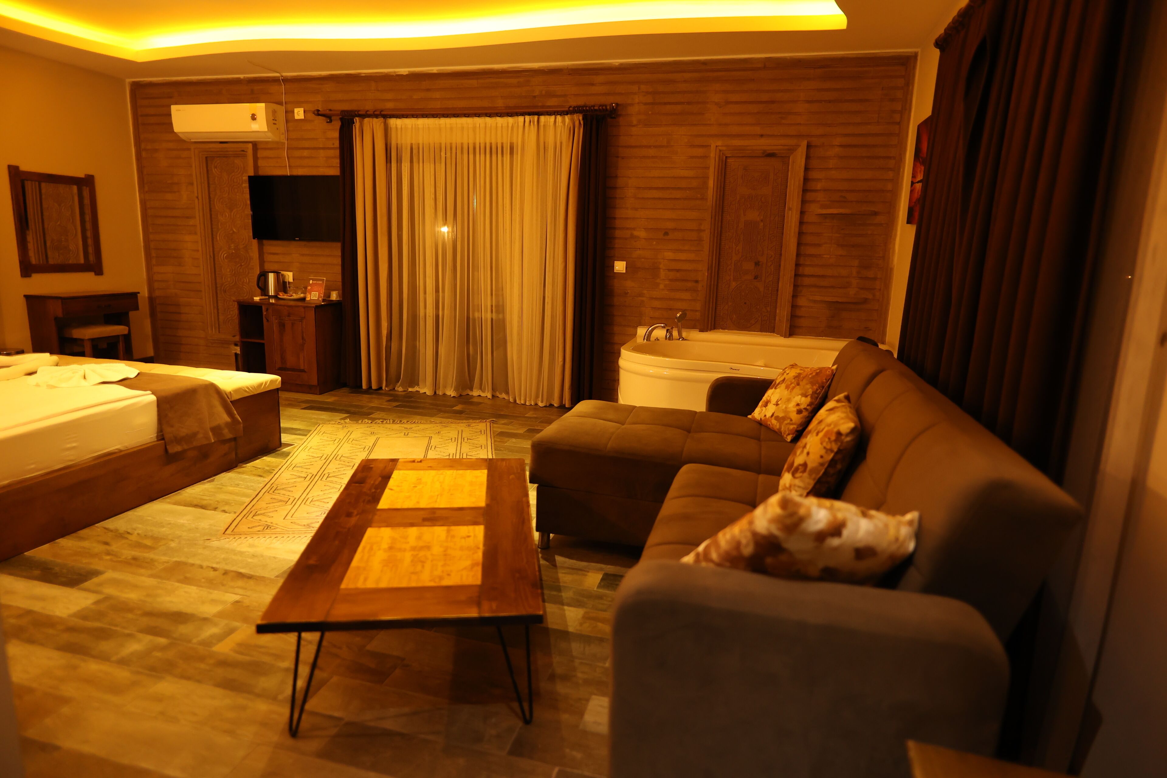 Una habitación de hotel moderna con sofá, una mesita de madera, cama, televisor y baño con bañera.