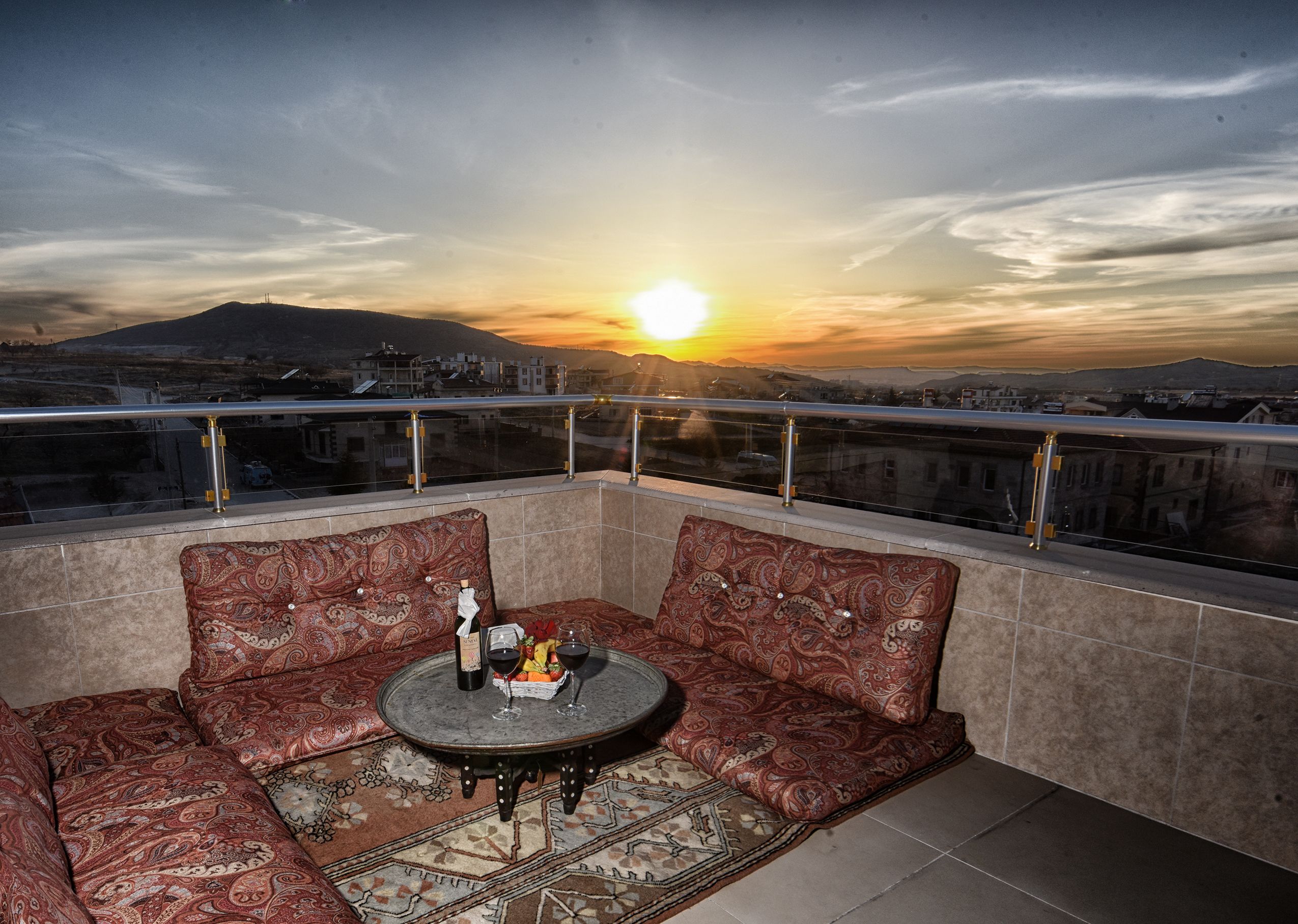 A balcony with a sofa and a small table, a sunset view, and a cityscape in the background.