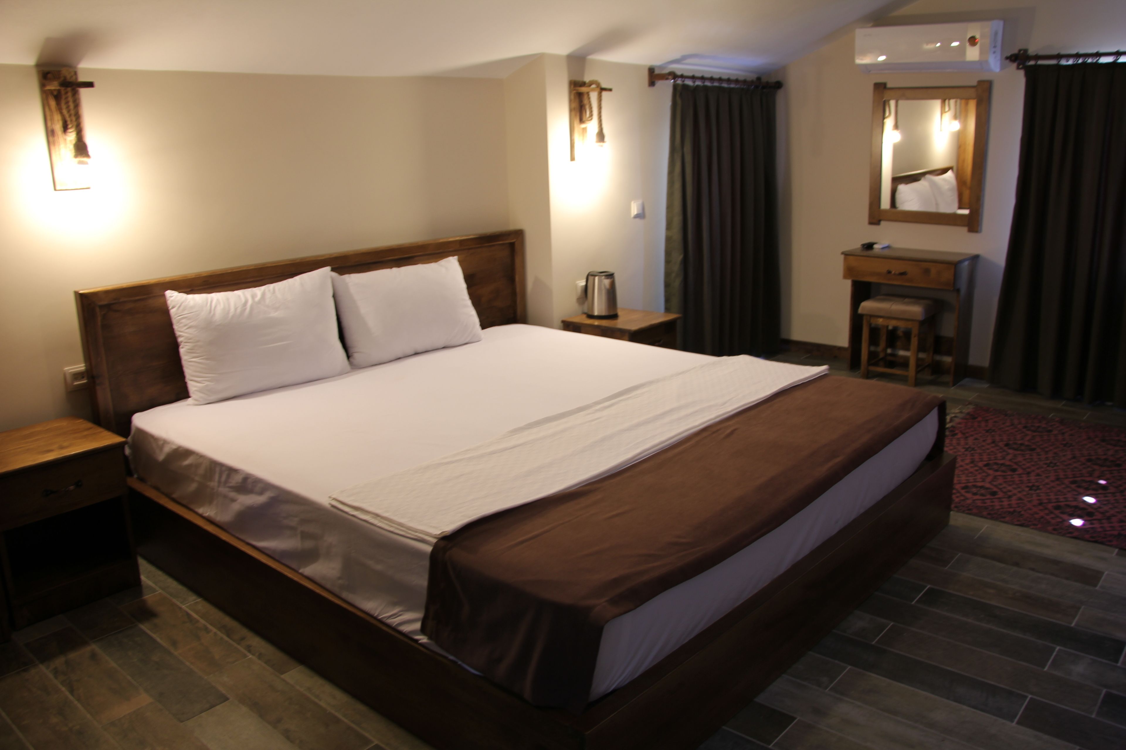 A hotel room with a large bed, bedside tables, a small desk with a chair, a mirror, and a red rug.