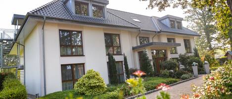 Une maison moderne à deux étages, dotée d’un toit en pente, de grandes fenêtres et d’un jardin bien entretenu où poussent diverses plantes et fleurs.