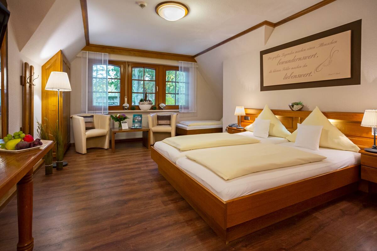 A hotel room with a large bed, a desk with a chair, a window with curtains, and a framed quote on the wall.