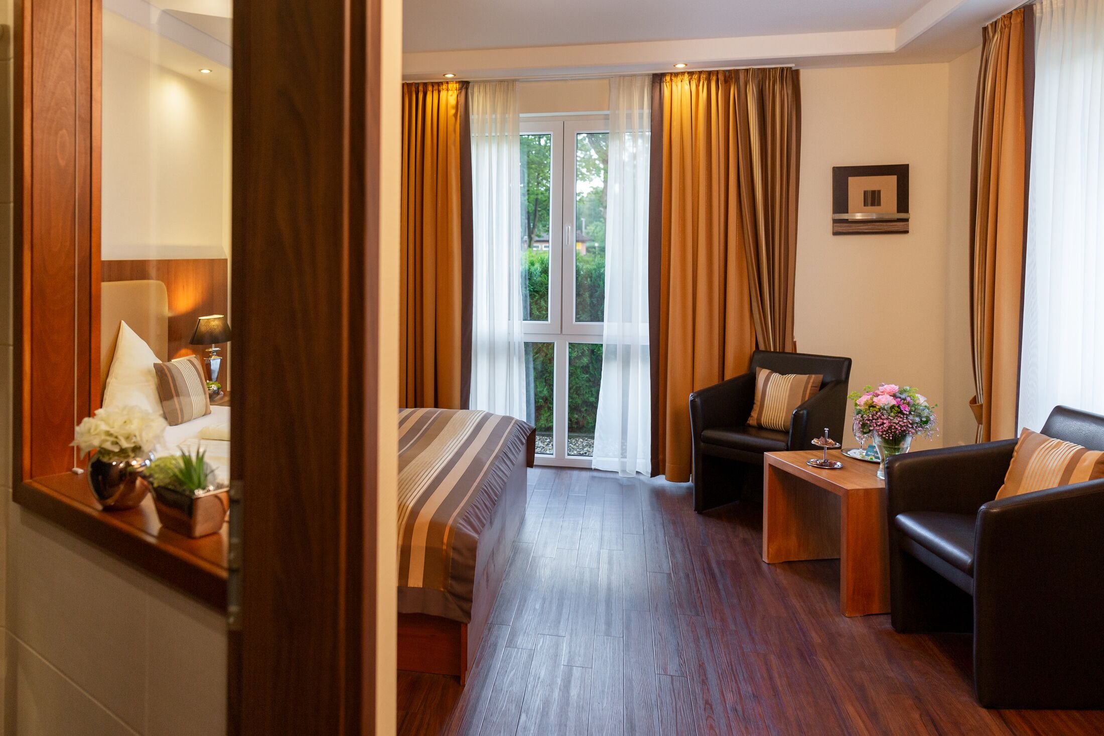 A hotel room with a bed, a sofa, a small table, and a chair. There is a window with curtains, and a door leading to another room.