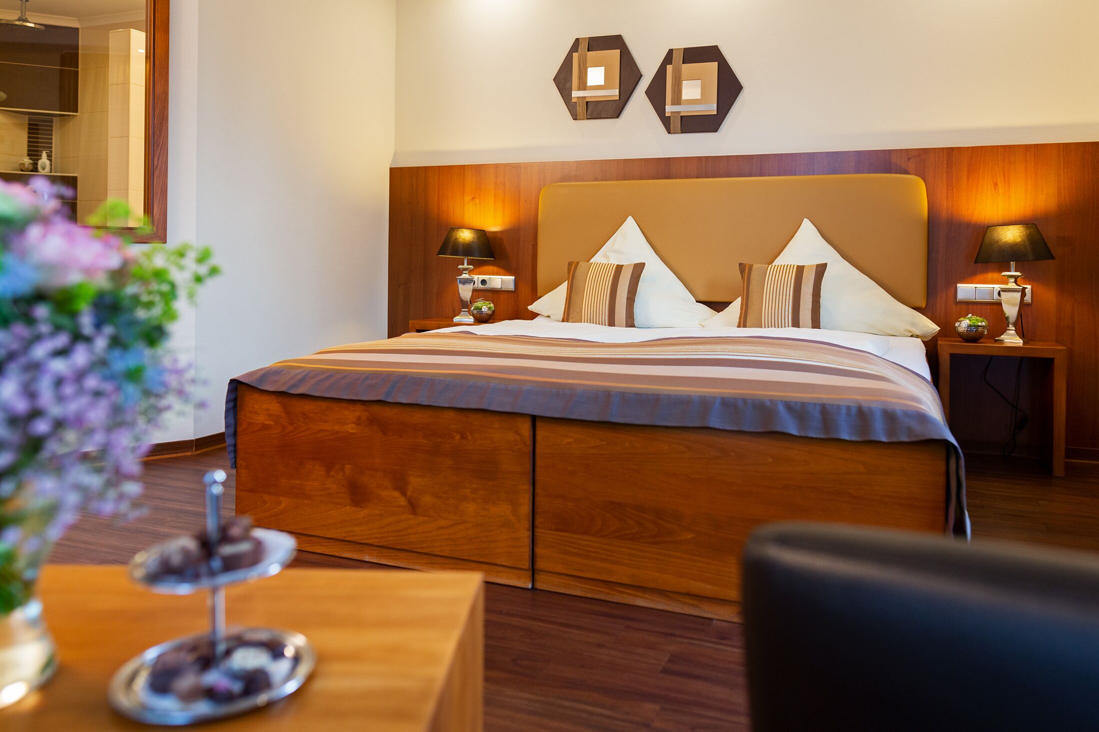 A hotel room with a large bed, wooden headboard, two bedside lamps, and a small table with a vase of flowers.