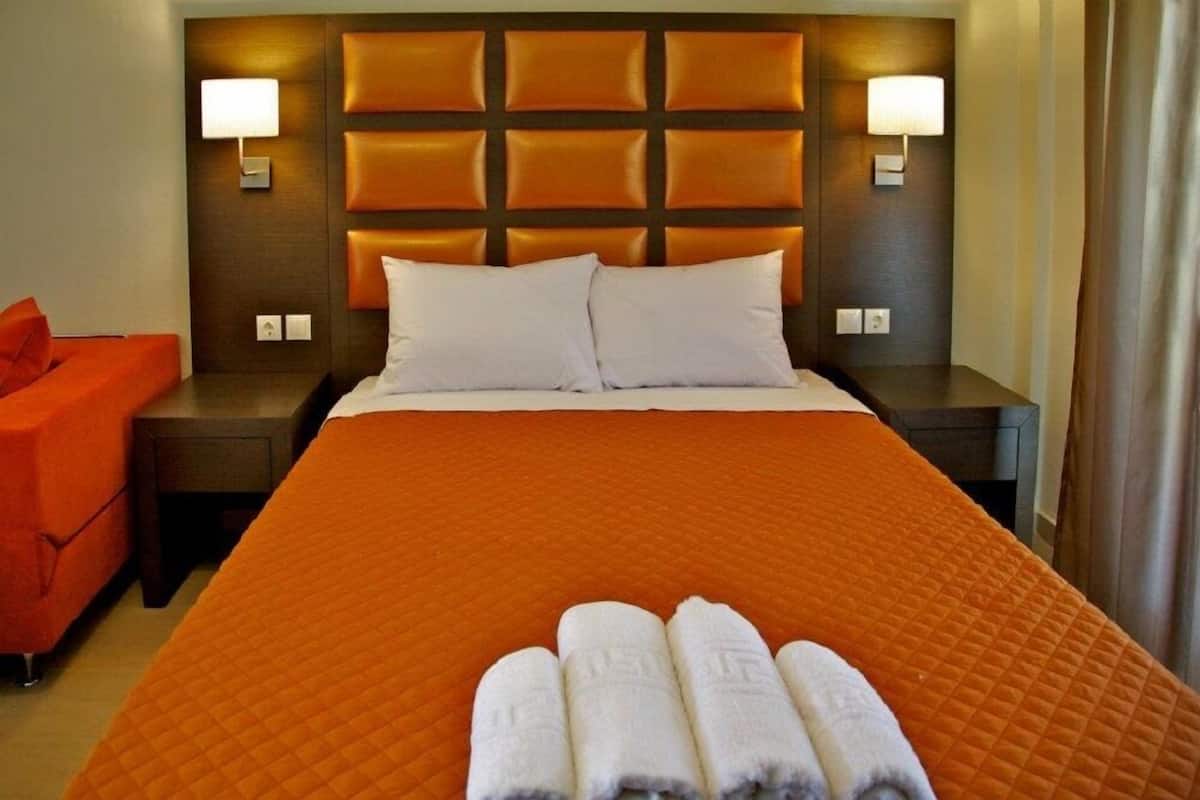 A hotel room with a large bed, two bedside tables, a nightstand, an orange chair, and a set of towels on the bed.