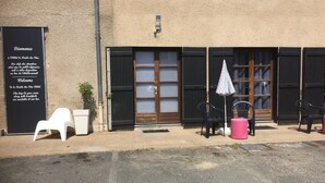 L’entrée d’un hôtel, avec une enseigne noire, une porte en bois et des sièges extérieurs composés de chaises en plastique et d’un tabouret rose.