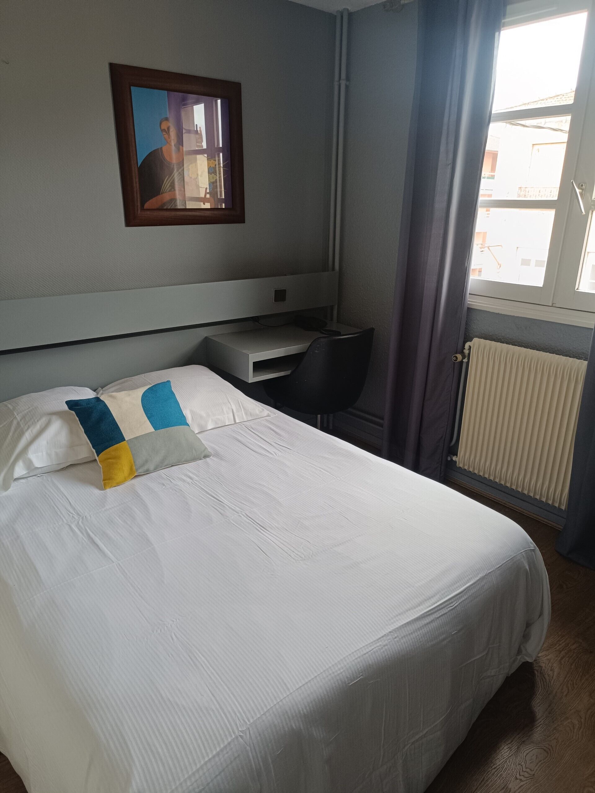 A hotel room with a bed, a desk, a chair, a radiator, and a window with curtains.