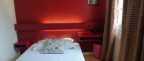 A bedroom with a red headboard, a bed with white bedding and a floral pillow, a bedside table, and a window with striped curtains.