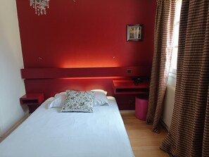 Une chambre à coucher avec une tête de lit rouge, un lit avec une literie blanche et un coussin à motifs floraux, une table de chevet et une fenêtre avec des rideaux à rayures.