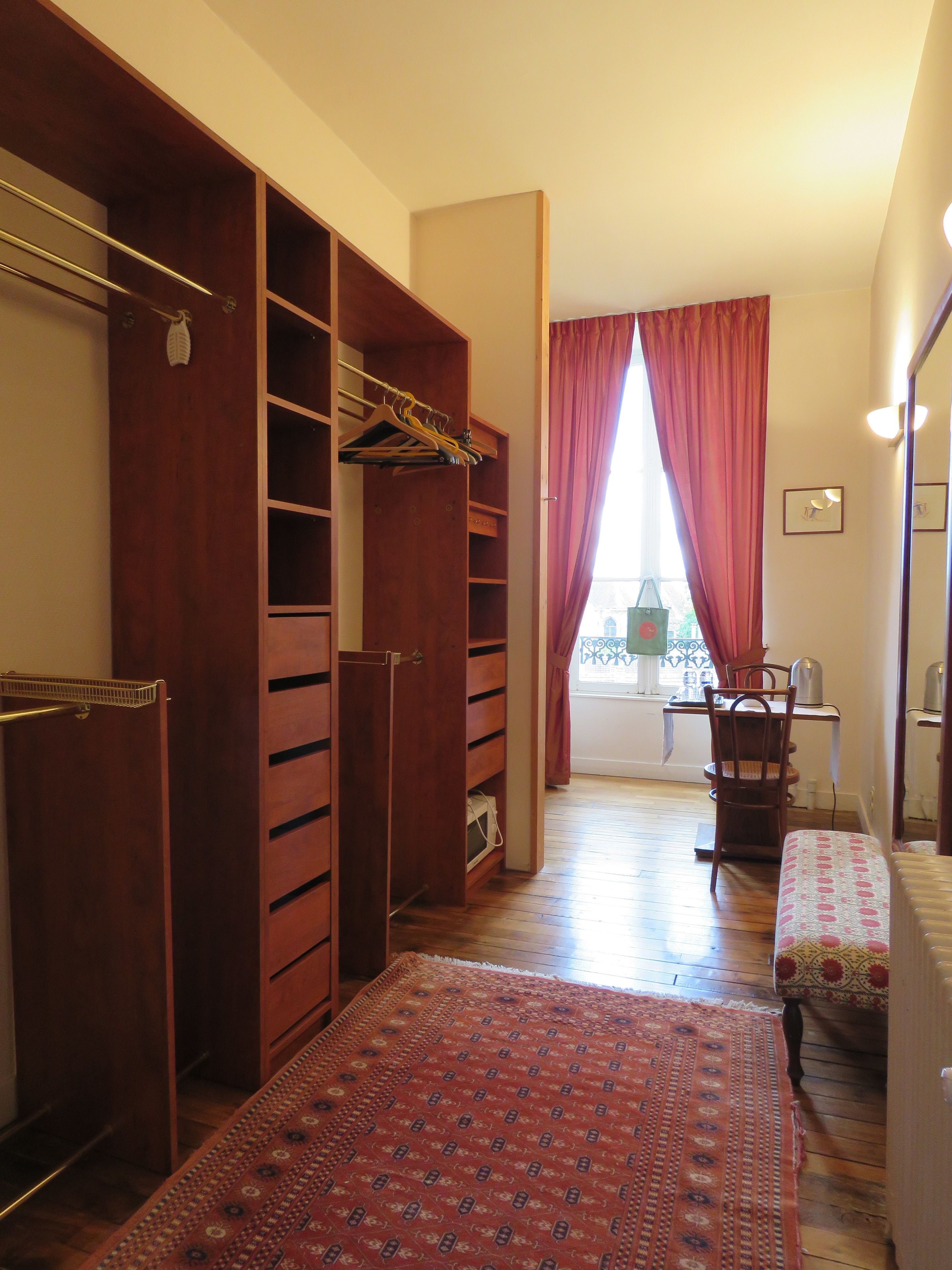 Una habitación con un armario de madera, un tapete estampado, una mesa de comedor con sillas y una ventana con cortinas.