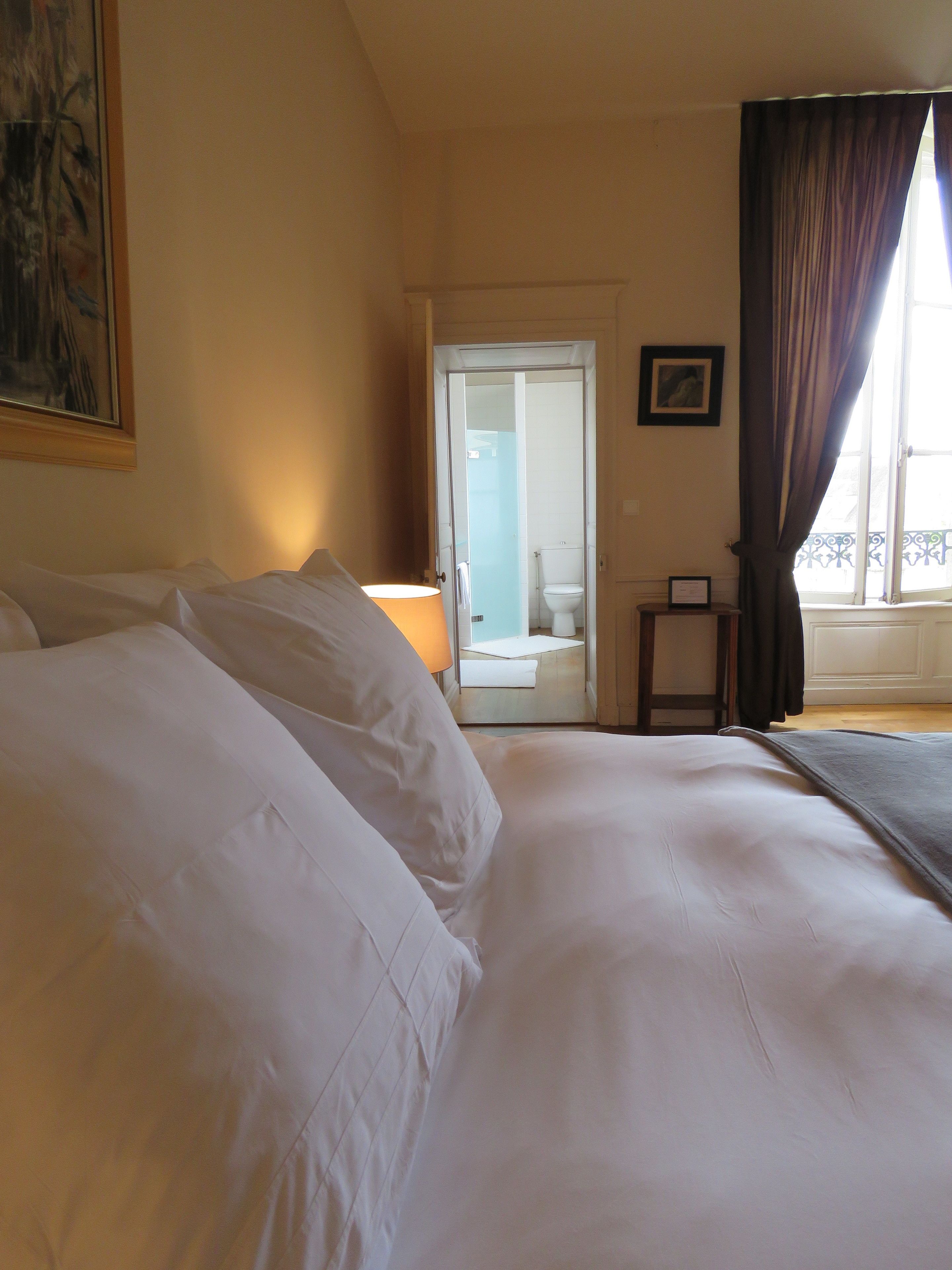 A hotel room with a large bed, a bedside table, a lamp, a window with curtains, and a door leading to a bathroom.