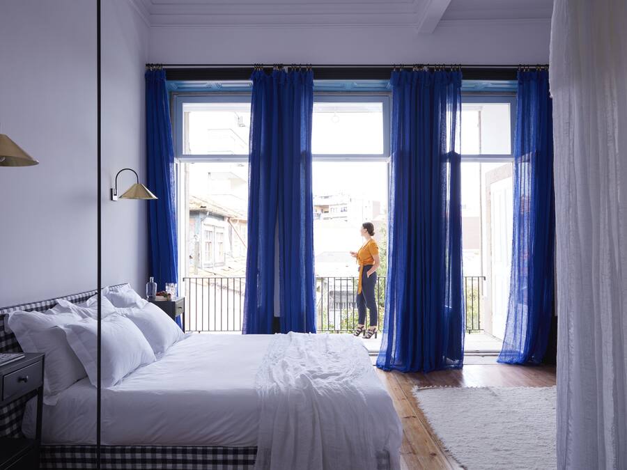 Un dormitorio con una cama grande, una mesita de noche, una lámpara y vistas a un balcón con cortinas azules.