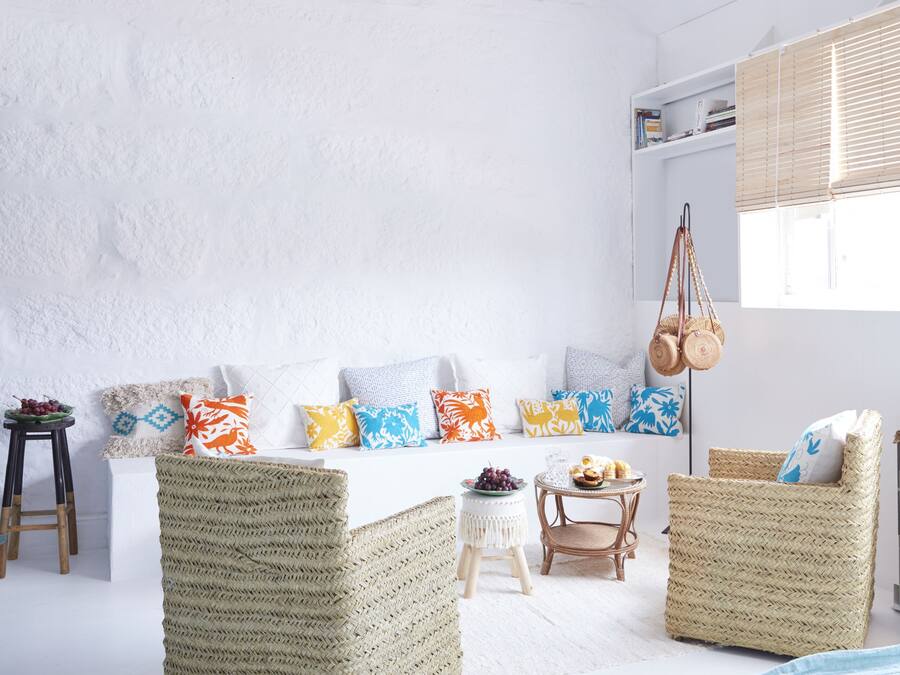 A room with a white wall, a woven sofa, a wooden chair, and a small table with a bowl of fruit.